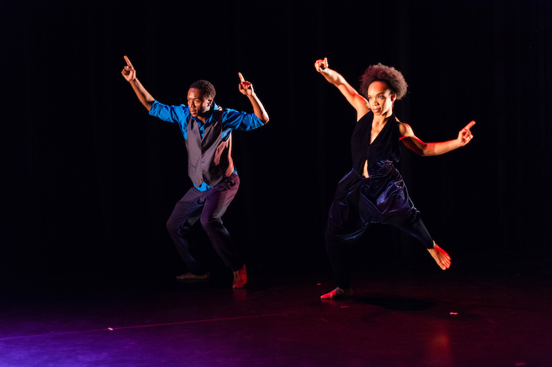 A female dancer in a long tunic juts out her right hip as her right leg hovers in the air at a 90 degree angle